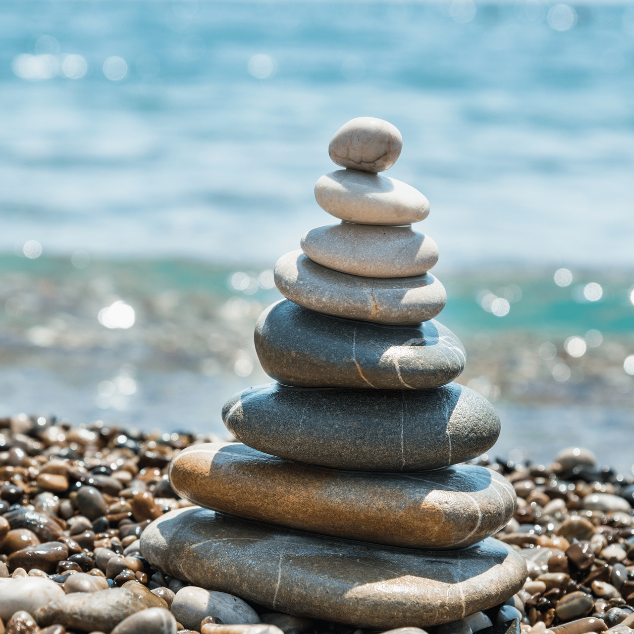 자연. 바닷가. 자갈밭 위에 쌓여있는 8층 돌탑. 그 뒤로 햇살에 반짝이는 푸른 바다.