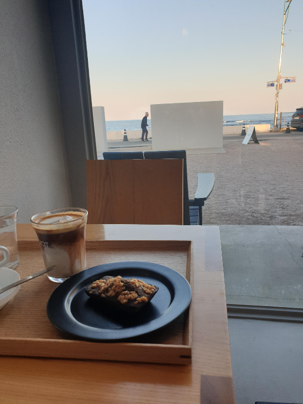 창문 너무 담장 사이 바다가 보인다. 내부 테이블에는 주문한 휘낭시에와 커피가 있다.