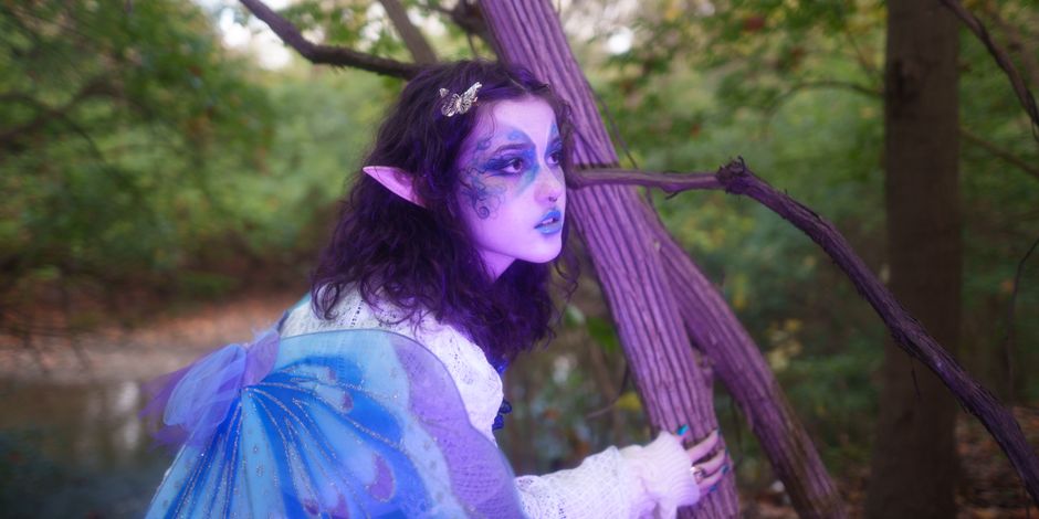 Enchanting elf with blue wings and intricate face makeup in a mystical forest setting.