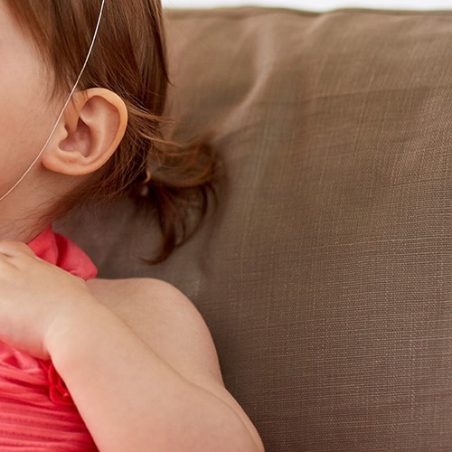 어린 아이의 옆모습의 일부가 보이며 아이는 갈색머리를 한쪽으로 묶었음. 배경에는 갈색 소파 또는 쿠션이 보여서 따뜻하고 부드러운 분위기를 담고있음.