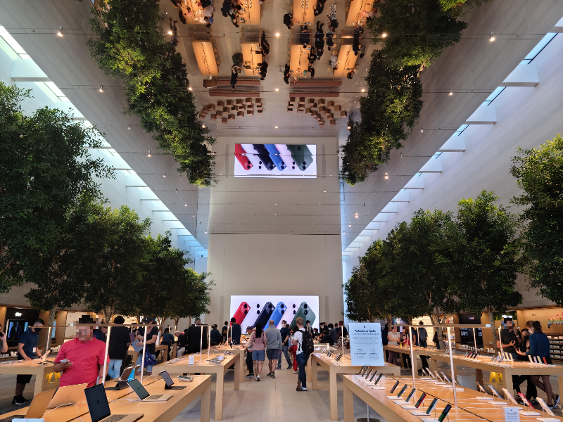 테이블이 가지런히 놓여 있고, 테이블 위에는 아이폰, 아이패드, 맥북 등이 놓여 있다. 앞쪽 벽에는 아이폰 광고가 나오고 있고, 양 옆으로는 실내에 커다란 나무들이 심겨 있다.