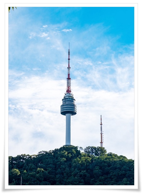 푸른하늘아래 산언덕 정상에 뽀족하고 우뚝 솟은 타워의 전경사진입니다. 타워의 중앙부에 사람들이 머물수 있는 건물층이 있어 보이고 아래쪽은 지상까지 굵고 둥근 기둥역활을 하고 중심부로부터 위쪽은 통신탑의 역할을 할것으로 보임