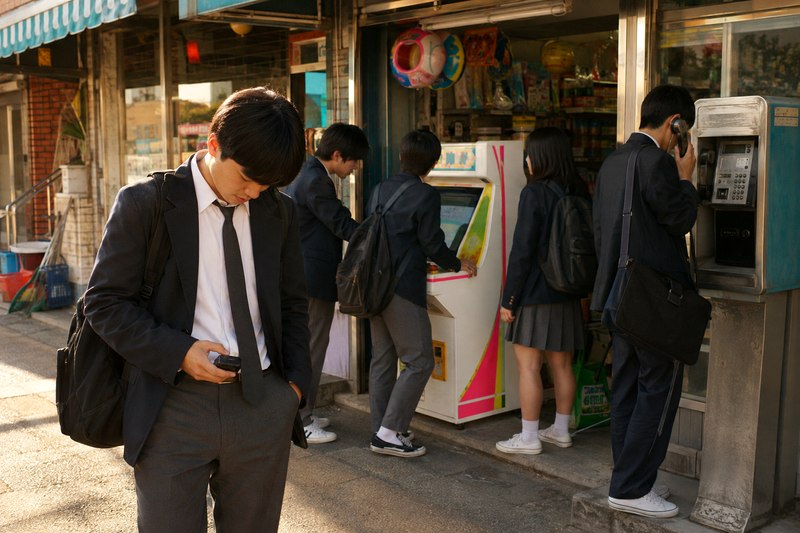 교복을 입은 학생이 문방구와 공중전화 앞에서 작은 삐삐 화면을 내려다보는 모습