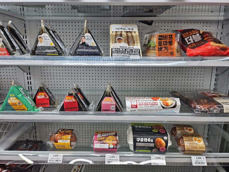 삼각김밥, 샌드위치, 햄버거, 훈제란 등 다양한 신선식품들이 진열된 편의점 냉장 매대의 전경. 유통기한이 짧아 매일 폐기 관리가 필요한 FF 품목들이 가득 찬 모습. 사장이 1%의 폐기율을 낮추기 위해 목숨을 걸어야 하는 동네 상권의 냉혹한 현실을 보여주는 진열대의 전경.