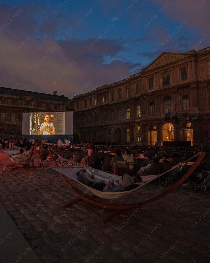 People relaxing on wooden hammocks inside the Cour Carree courtyard of the Louvre Museum watching a movie on a large outdoor screen at dusk