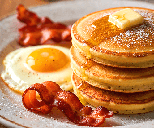 하얀 접시에 3단 팬케이크가 있고 옆에는 계란프라이와 구운 베이컨이 있다.