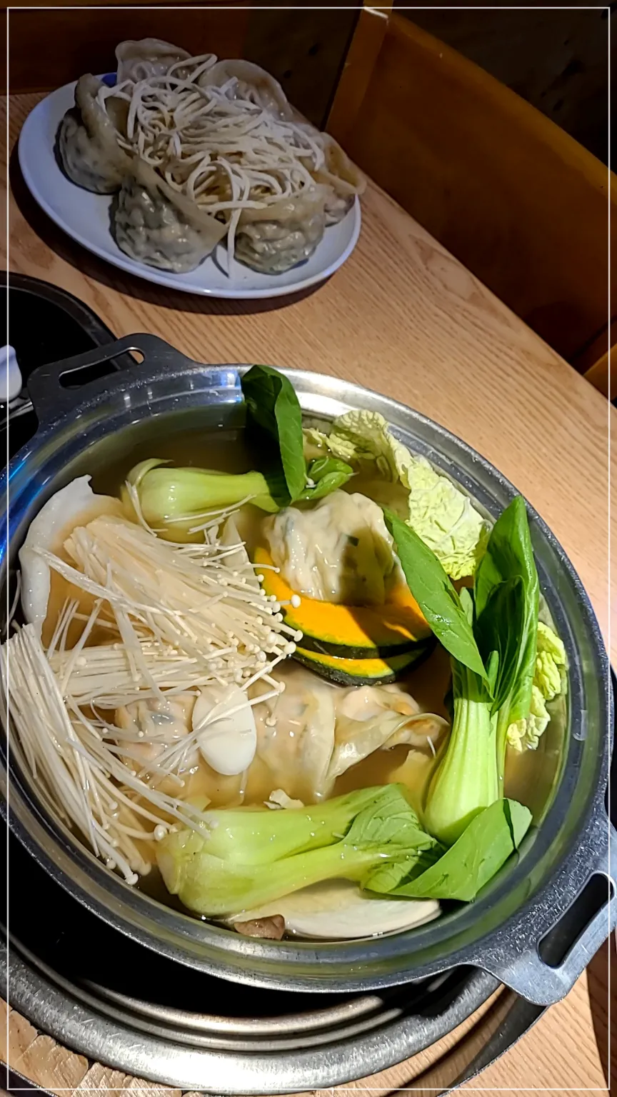 테이블위에 만두전골냄비 청경채 단호박 버섯 만두가 들어 있음. 흰접시에는 만두6개와 칼국수가 놓여있음
