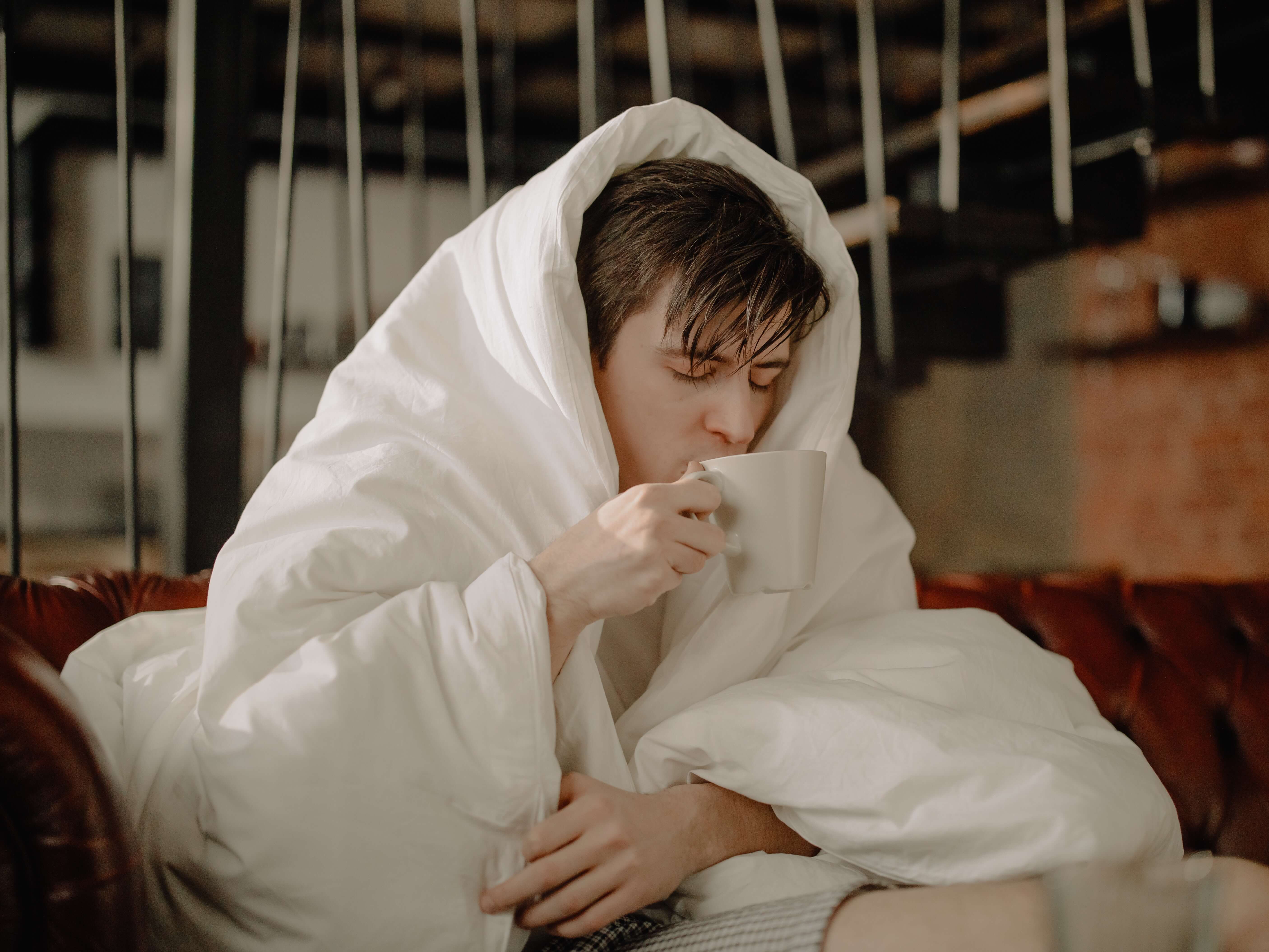 독감에 걸린 남성이 이불을 뒤집어 쓰고 따뜻한 차를 마시는 장면