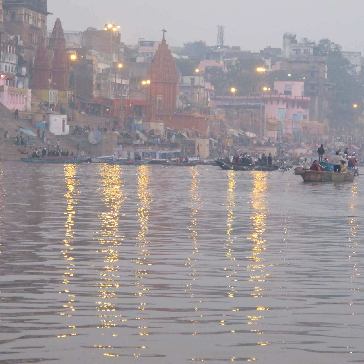 이른 새벽 안개가 자욱한 갠지스강 위로 여러 척의 보트가 떠 있고, 강물에 도시의 불빛이 황금빛으로 반사되는 풍경
