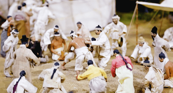 상투를 한 남자들이 씨름판에서 씨름을 하고, 구경중이다.