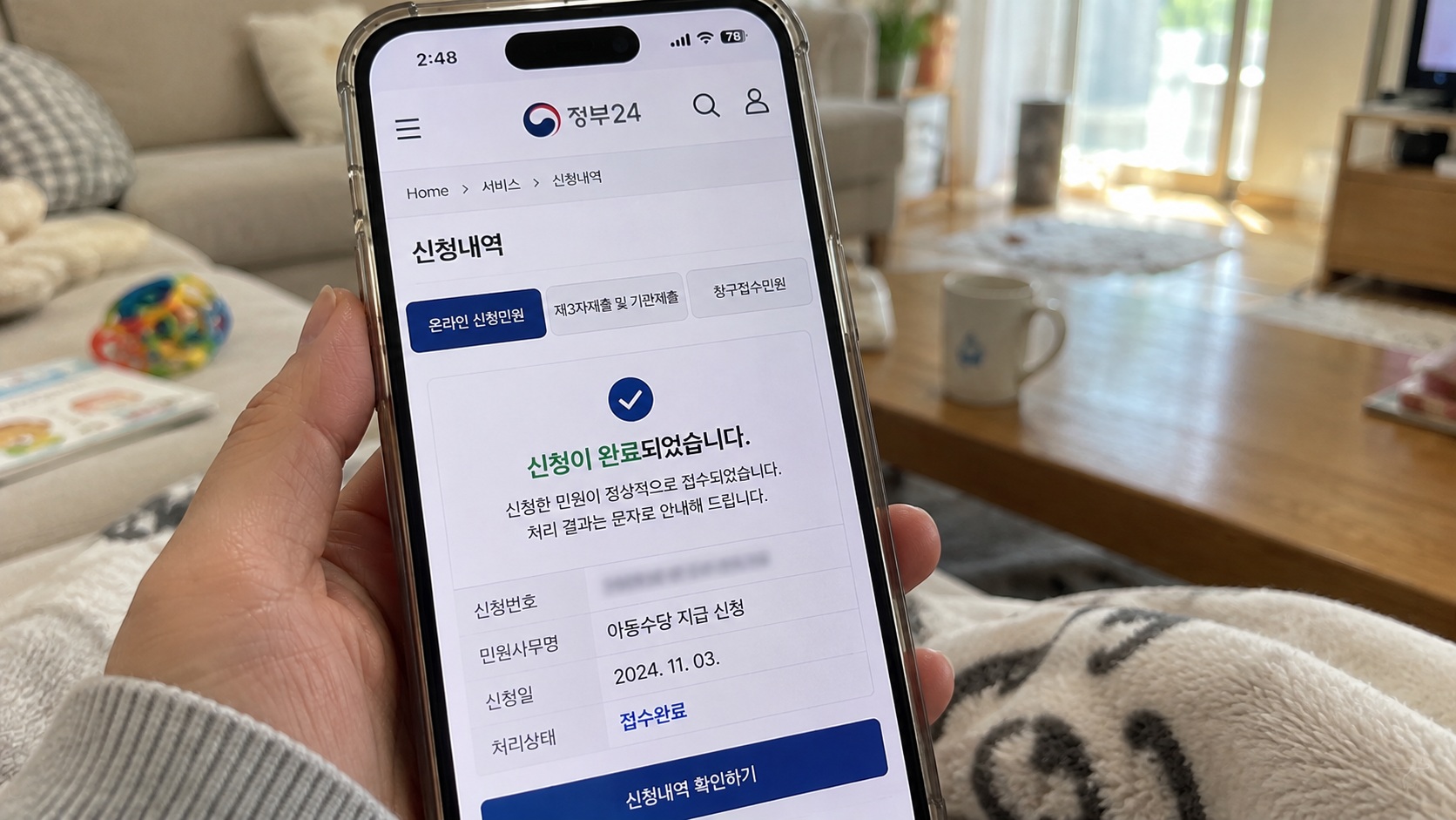 거실에서 스마트폰으로 정부24 아동수당 지급 신청 완료 화면과 접수 상태를 확인하는 모습
