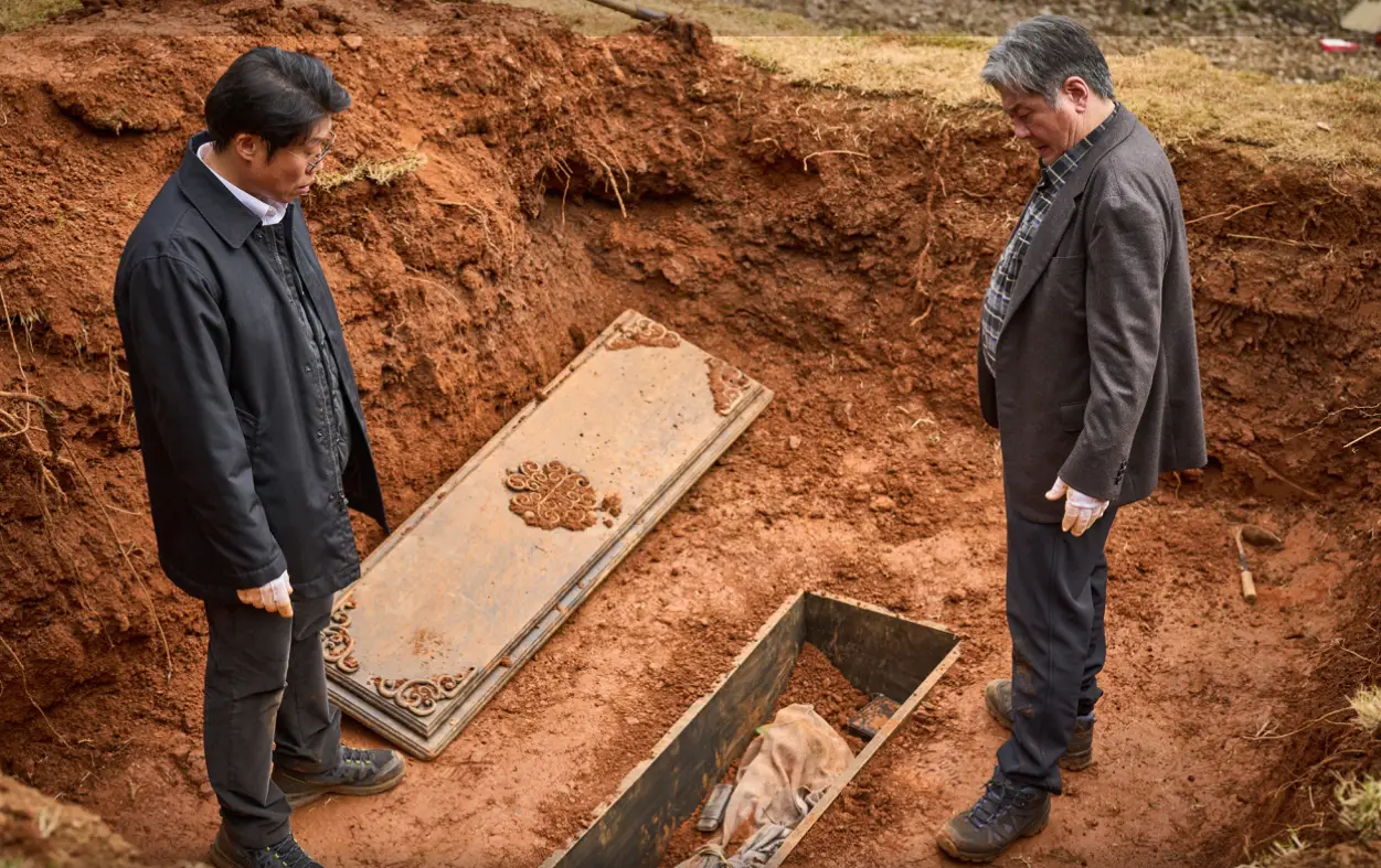 파묘- 갈색 흙이 파져있는 가운데 뚜껑 열린 관과 그안에 흰 천으로 덮여진 것을 바라보는 좌측 유해진과 우측 최민식