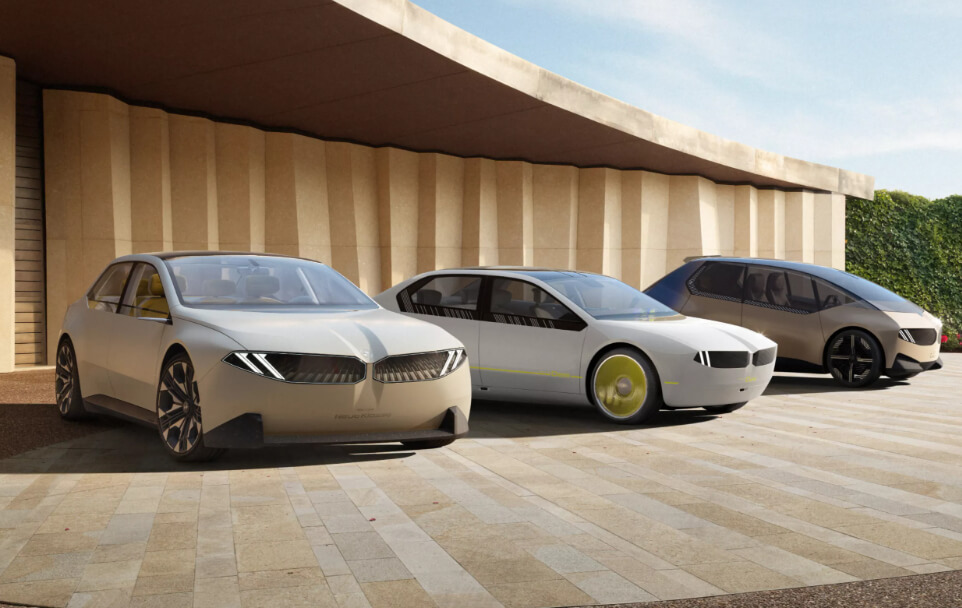 미래지향적 디자인의 BMW Neue Klasse 전기차 3대가 현대적인 건축 구조물 앞에 주차되어 있습니다. 차량은 깔끔한 외관과 BMW 특유의 조명이 들어온 그릴이 돋보입니다.