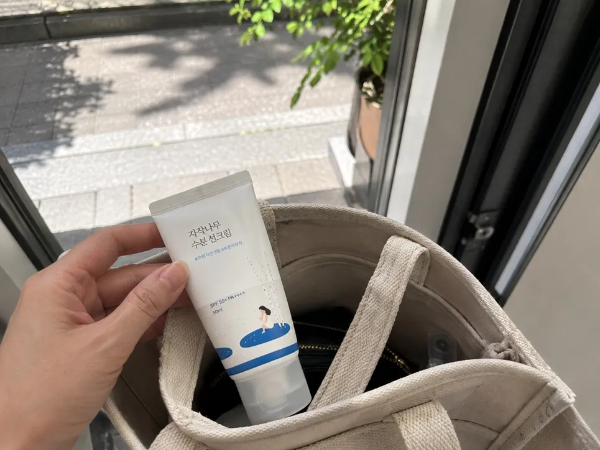 야외 보도블록과 초록색 나뭇잎이 보이는 밝은 문앞에서, 한 손으로 베이지색 캔버스 에코백 가방 안에 라운드랩 브랜드의 '자작나무 수분 선크림' 튜브형 제품을 집어넣거나 챙기고 있는 일상적인 모습을 촬영한 사진