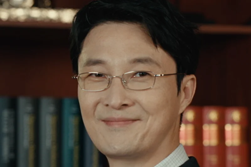 어느 서재를 배경으로 검은 정장을 입고 금테안경을 쓴 채 웃고 있는 나상우