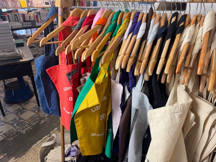 Colorful canvas tote bags with Ofr typography hanging on wooden hangers inside Ofr bookshop Paris ❘ Tote bag Ofr Paris