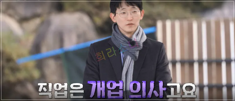 광수가 야외에서 서서 인터뷰를 하는 모습이다. 그는 검은색 코트에 회색 목도리를 두르고 있으며&amp;#44; 얇은 금속 테 안경을 착용하고 있다. 차분한 표정으로 화면을 바라보고 있다. 자막: 