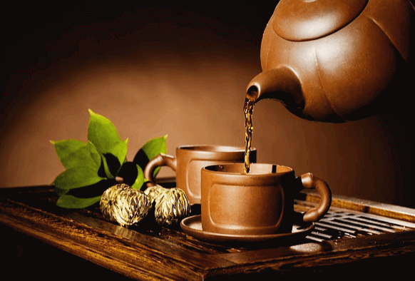 갈색 도자기 주전자에서 녹차물이 찻잔에 떨어진다. 식탁엔 2개의 갈색 찻잔이 있다.