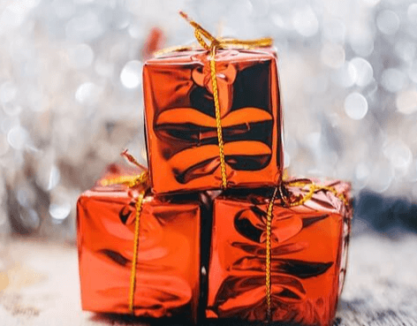 번쩍거리는 주황빛 포장지로 포장된 작은 선물상자가 3개 쌓여있고 뒷 배경은 불빛에 반짝거린다.