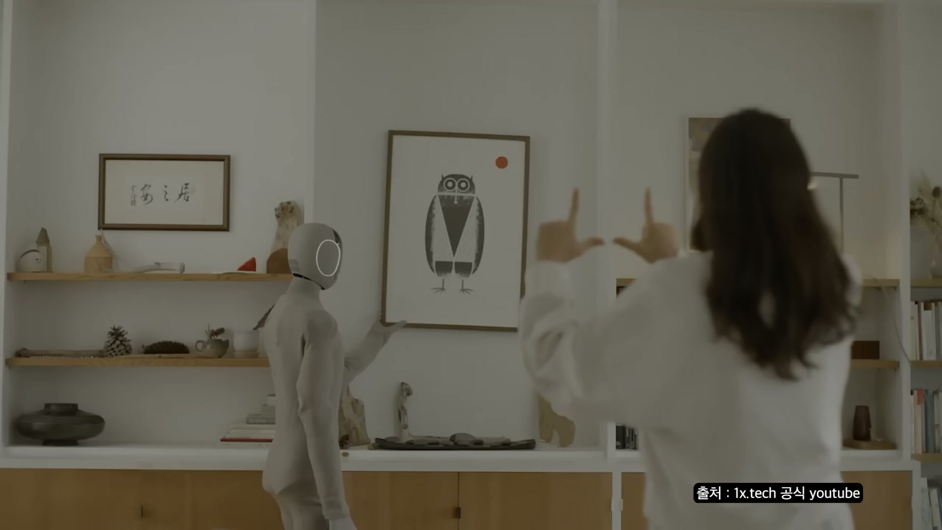 흰 벽에 액자가 걸린 거실에서 회색 휴머노이드 로봇이 한 여성과 함께 손을 들고 가리키는 동작을 하고 있다.