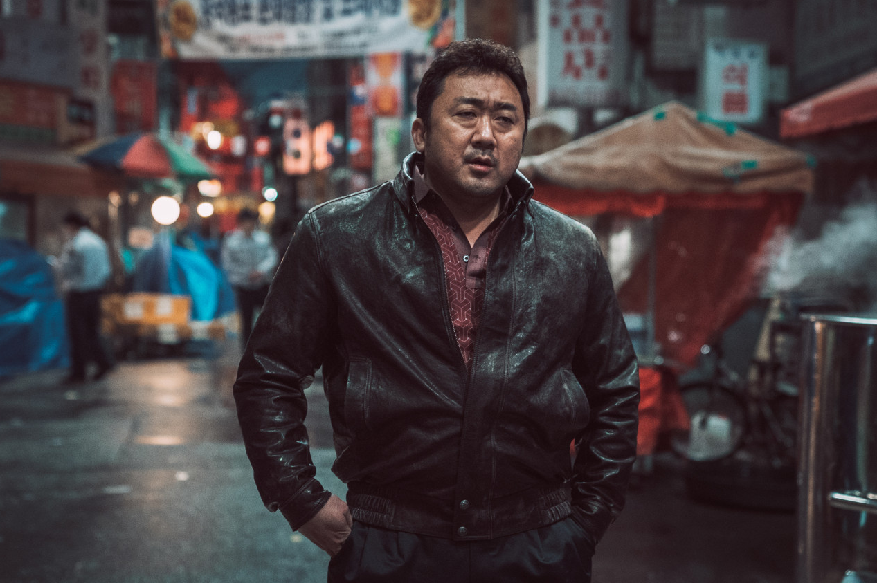 밤거리 네온사인이 가득한 가리봉동 골목에서 마석도가 두 손을 주머니에 넣고 정면을 바라보며 홀로 서 있는 장면