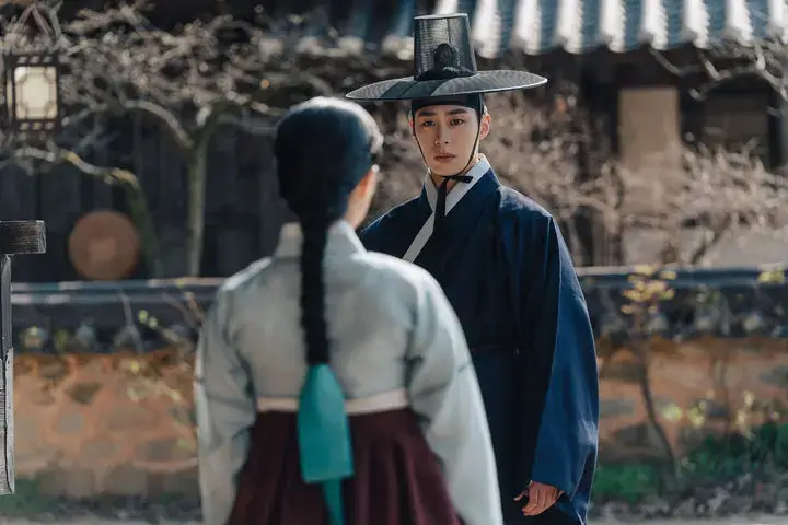 《탄금》의 한 장면. 전통 한옥 마당에서 푸른 도포를 입은 홍랑이 정면을 응시하고 있고, 등을 보인 재이가 그를 마주하고 서 있다. 두 인물 사이에 긴장감이 흐르는 순간이 포착되어 있다.