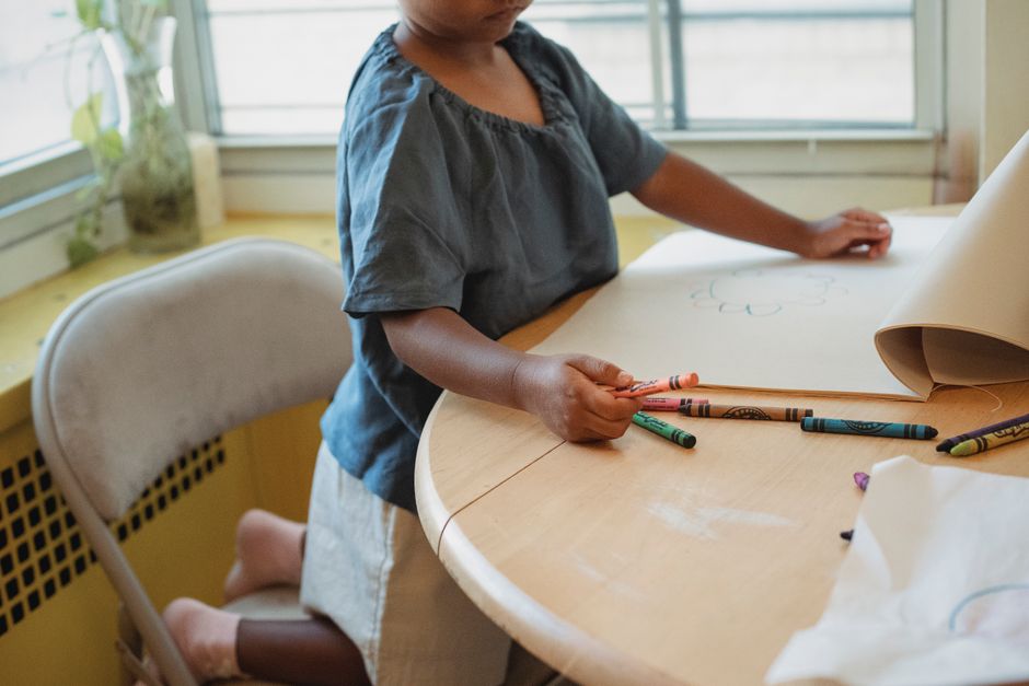 Crop anonymous child in casual outfit with color wax crayons and paper sitting at table and drawing