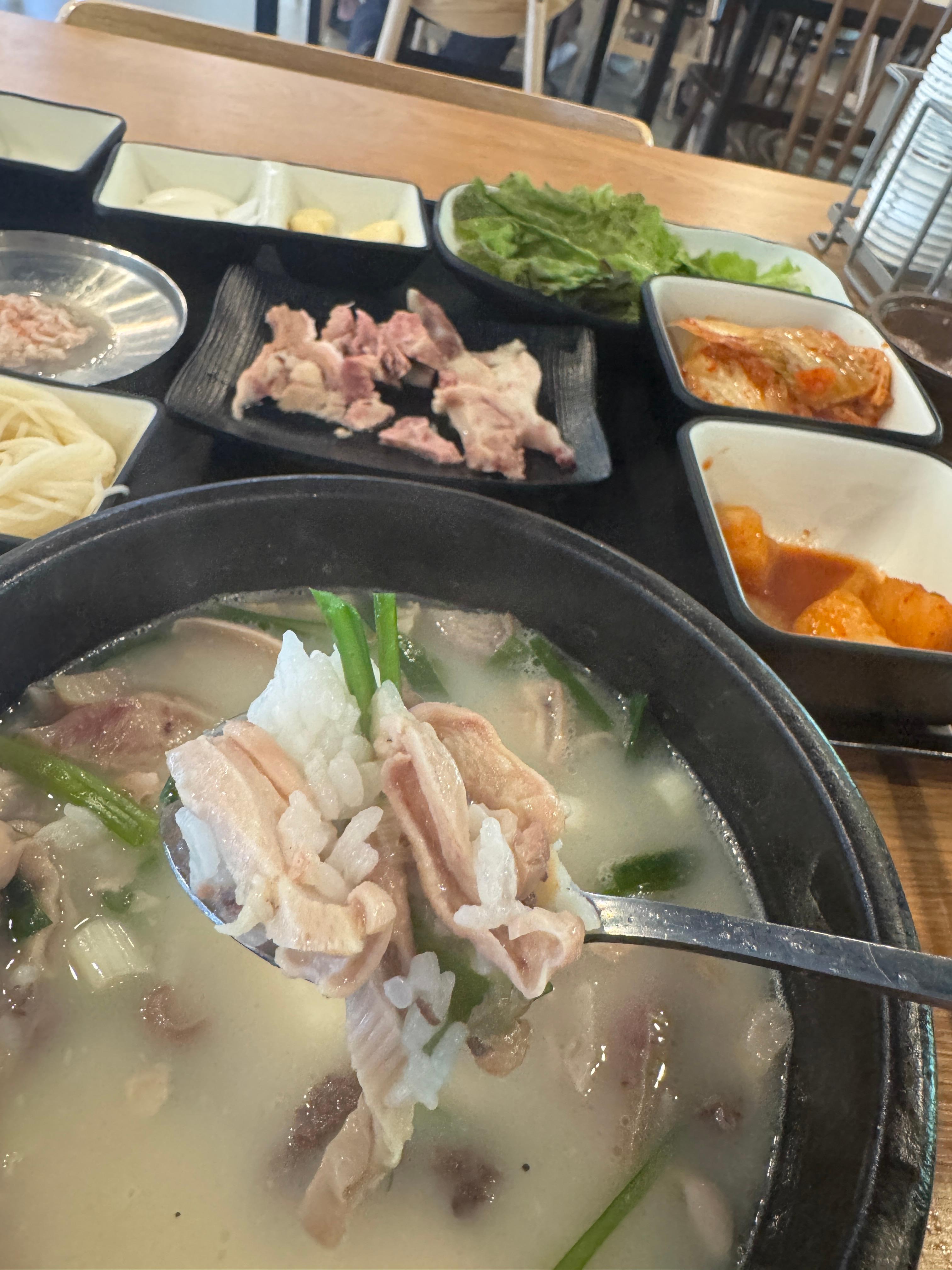 뚝배기 속 국밥을 숟가락으로 크게 떠내는 장면. 김치&amp;#44; 깍두기&amp;#44; 수육&amp;#44; 소면 등 밑반찬이 테이블 위에 놓여 있다.