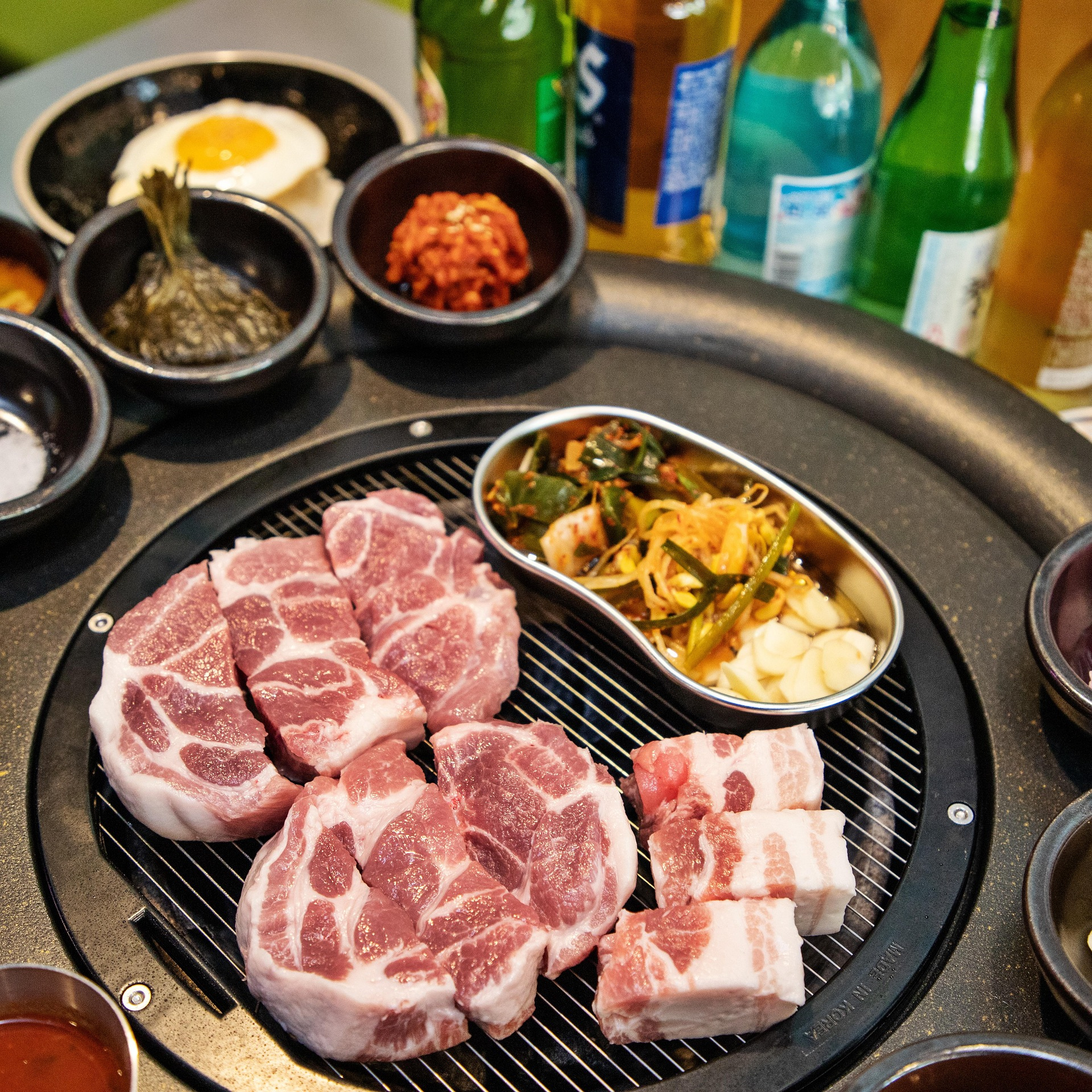 Fresh cuts of samgyeopsal (Korean pork belly) sizzling on a tabletop grill surrounded by side dishes, garlic, kimchi, and bottles of soju &mdash; a classic Korean BBQ scene that captures the communal warmth and flavor of Korean dining culture.