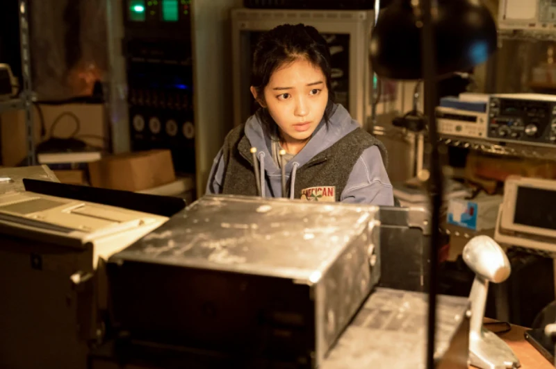 허름한 기기들을 배경으로 후드티를 입고 모니터를 바라보고 있는 영화 더 문의 한별