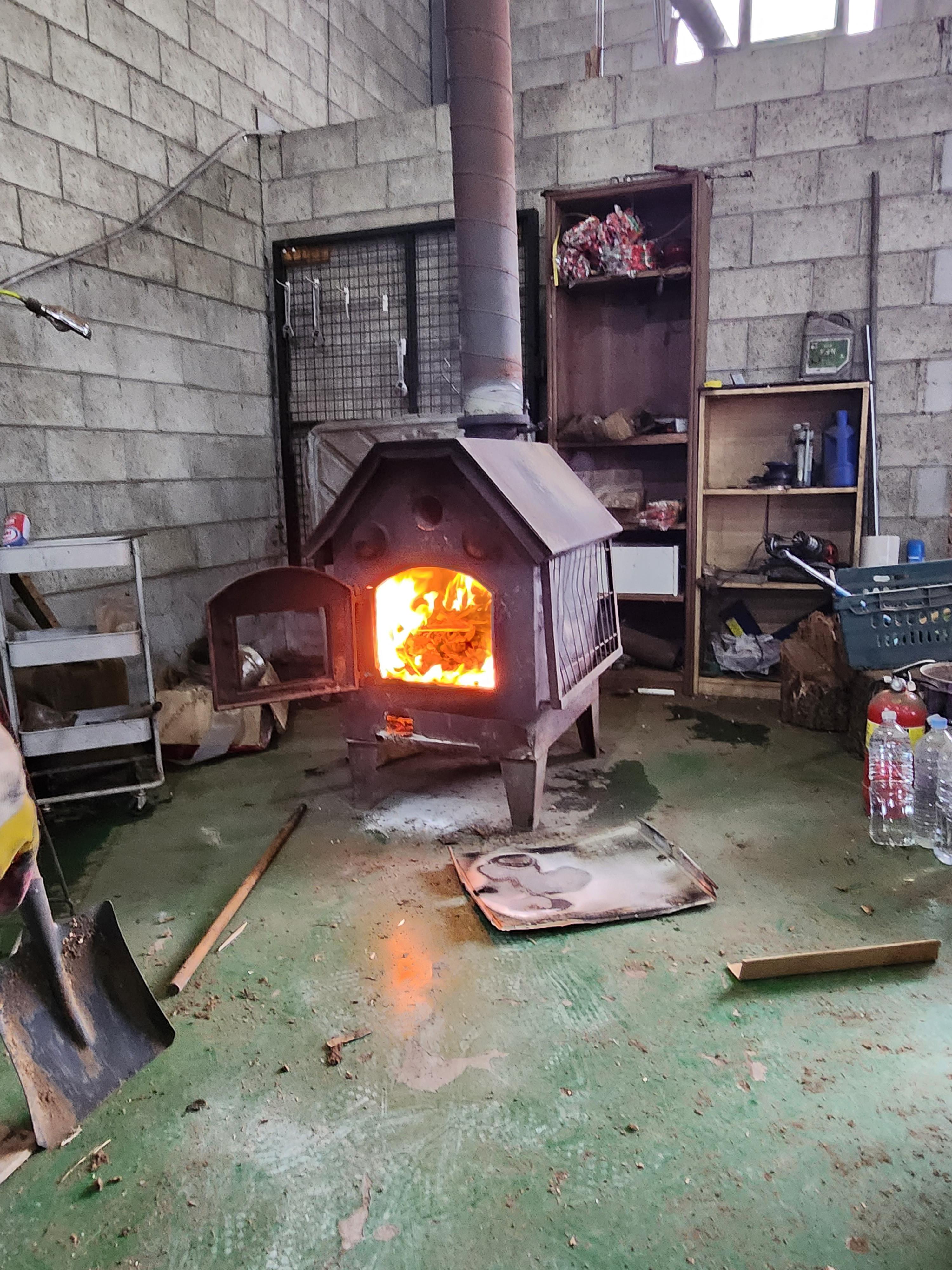 콘크리트 벽돌 벽으로 마감된 어수선한 작업실 내부에 주황색 불꽃이 활활 타오르는 검은색 금속 난로가 놓여 있다. 난로 앞 바닥은 젖어 있고 타다 남은 재가 흩어져 있다. 난로 뒤편으로는 목재 선반과 금속 철망이 보이고, 왼쪽으로는 흰색 트롤리와 삽이 놓여 있다.
