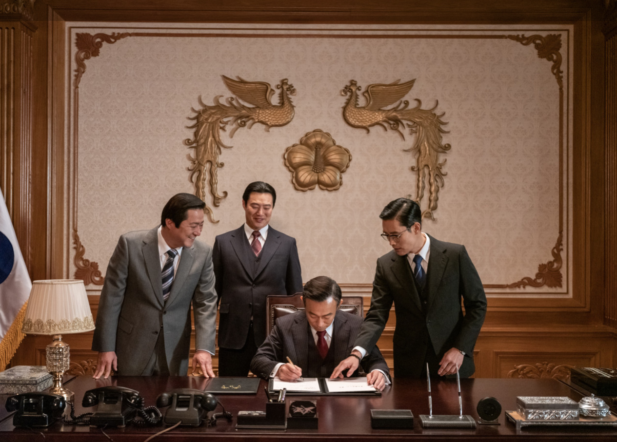 화려한 봉황 문양이 장식된 청와대 집무실에서 대통령이 서류에 서명하는 가운데 김규평을 포함한 측근들이 주변에 서 있는 장면
