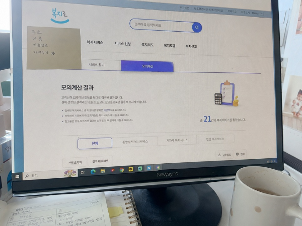 복지로 홈페이지의 모의계산 결과 화면이 켜진 컴퓨터 모니터와 그 앞에 놓인 육아 기록 수첩 및 머그잔의 모습