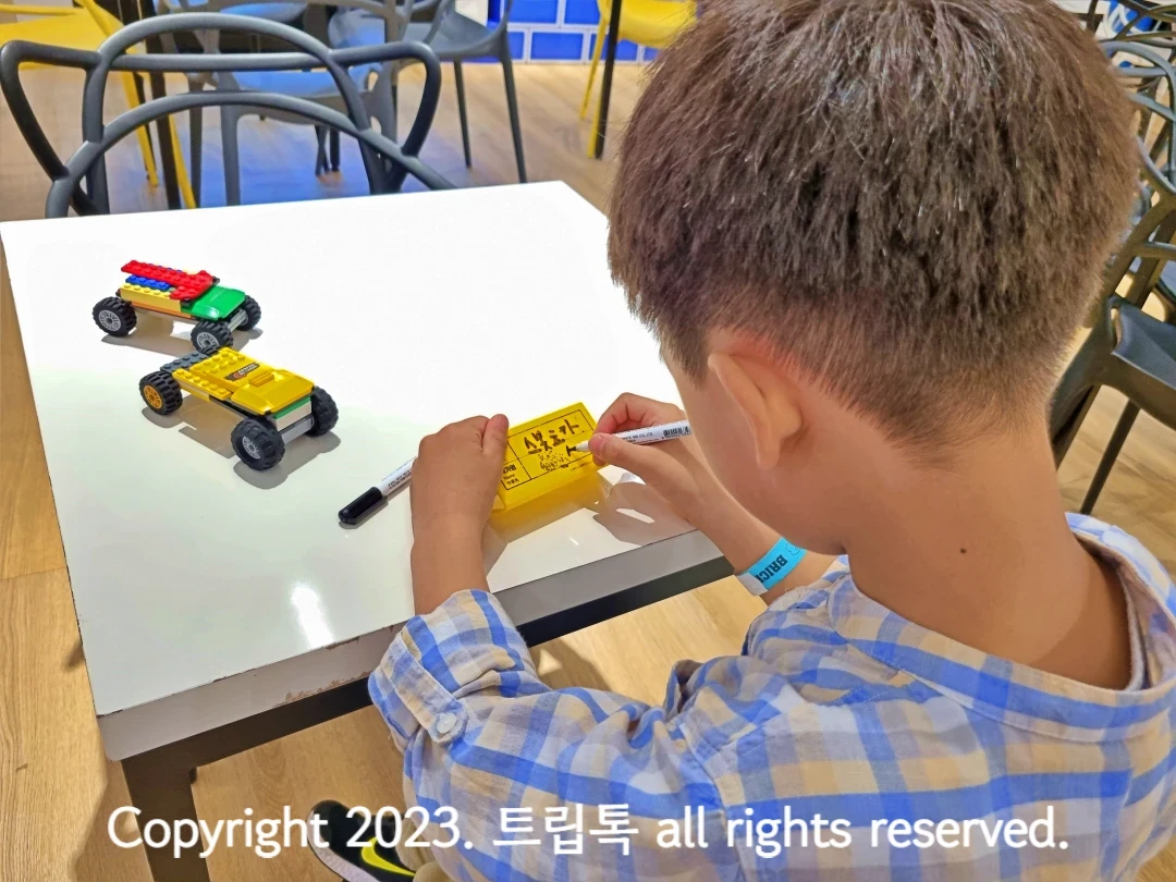 왼쪽에 브릭으로 만든 자동차2대가 있고 노랑 명찰에 작품이름과 아이이름을 적는 뒷모습
