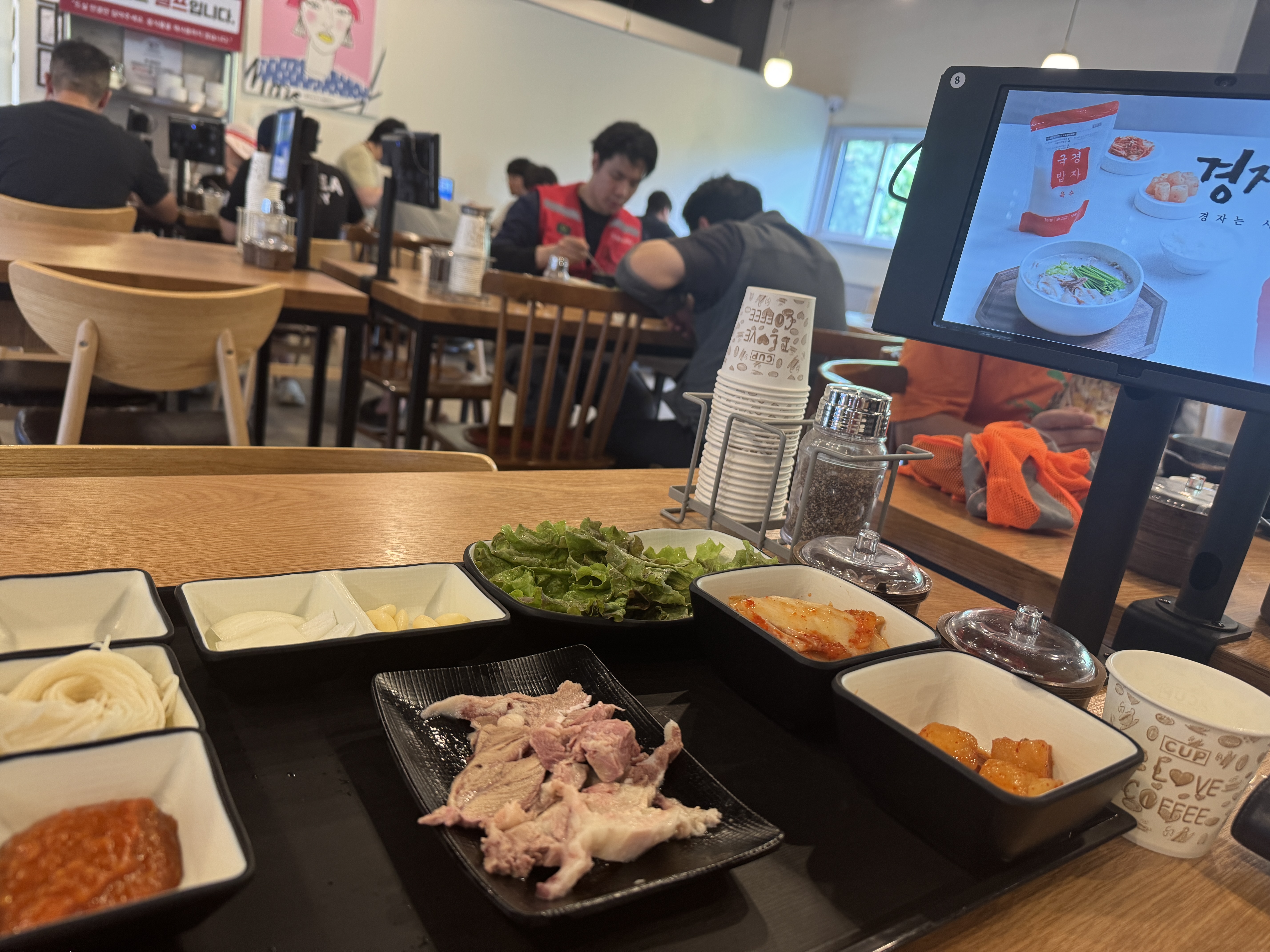 경자국밥 용인본점 실내 테이블 전경. 작은 접시에 담긴 수육과 함께 소면, 쌈채소, 양파&middot;마늘, 김치, 깍두기, 쌈장 등 밑반찬이 차려져 있다. 테이블 옆에는 키오스크 화면이 보인다.
