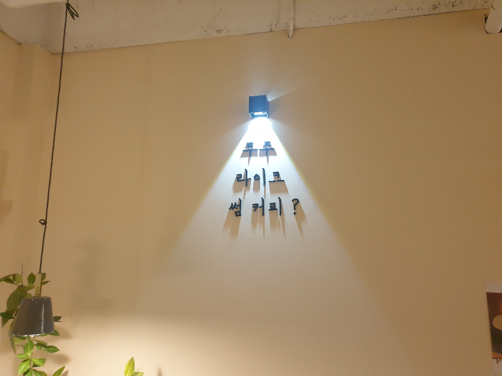 벽에 글자가 부착되어 있다. 무주 라이크 썸 커피? 라는 글자가 있고 그 글자에 조명이 빛을 비추고 있다.