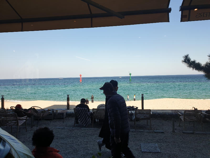 창문 밖의 풍경.바다와 바다를 바라보고 앉는 테이블과 의자. 유리 앞을 지나는 사람들