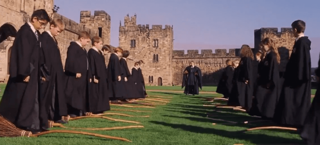 Magic school freshmen are standing next to broomsticks. This is the first broom flying class.