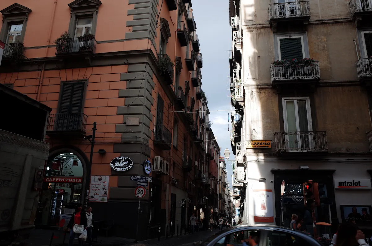 나폴리의 좁고 높은 건물들 사이 골목길 풍경, 1층에 위치한 Caffè del Professore 간판과 거리의 사람들