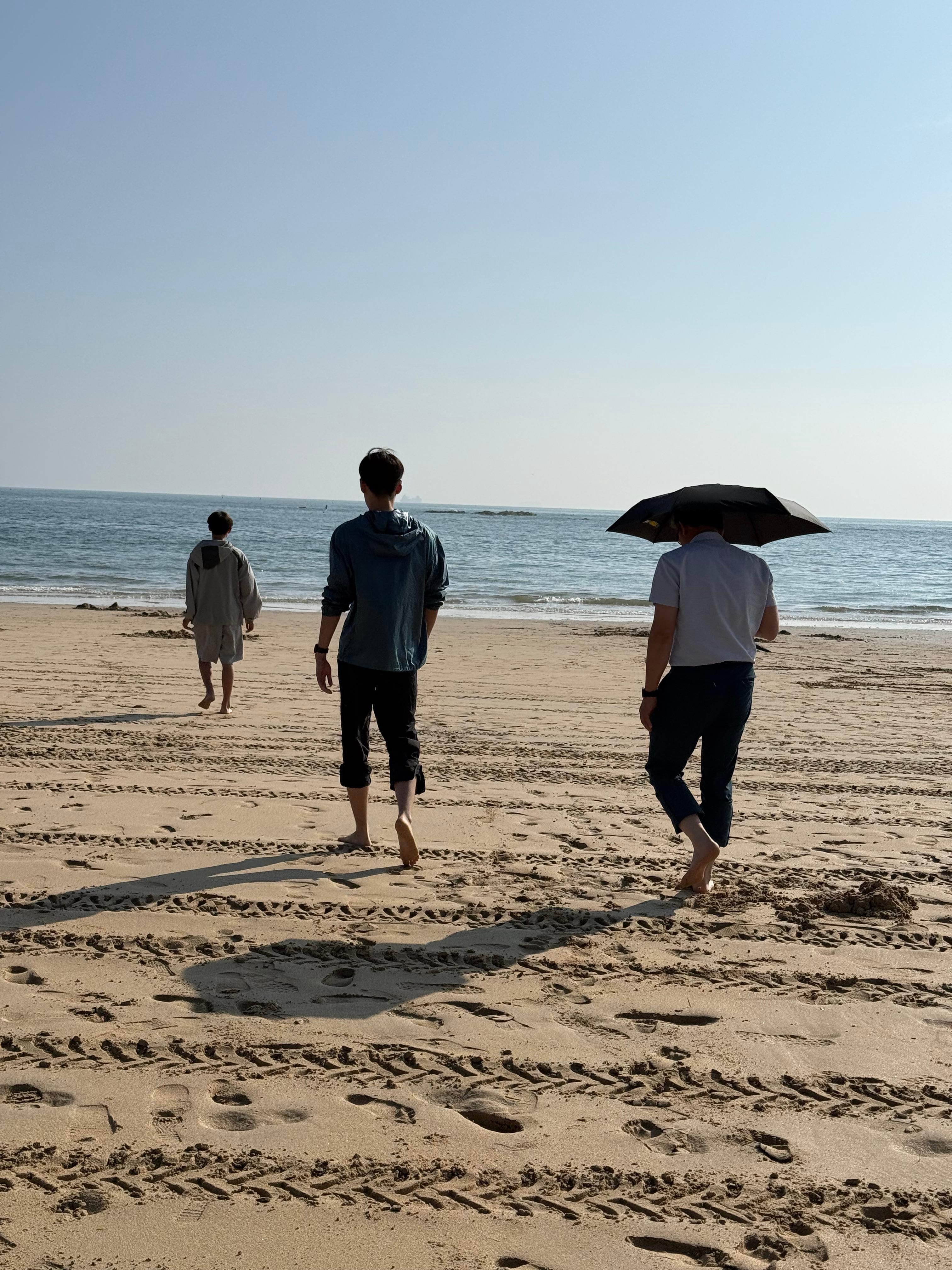 지인들과 해변가를 향해 맨발로 걸어가는 모습입니다. 한 사람은 검은 우산을 쓰고 햇빛을 피하고 있고 두 사람은 햇빛을 맞으며 걸어가고 있습니다.