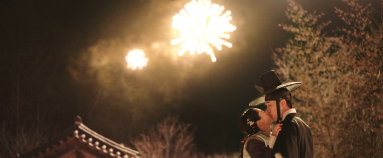 밤하늘에 불꽃놀이가 펼쳐지는 가운데, 한복을 입은 남녀가 로맨틱한 키스를 나누는 장면