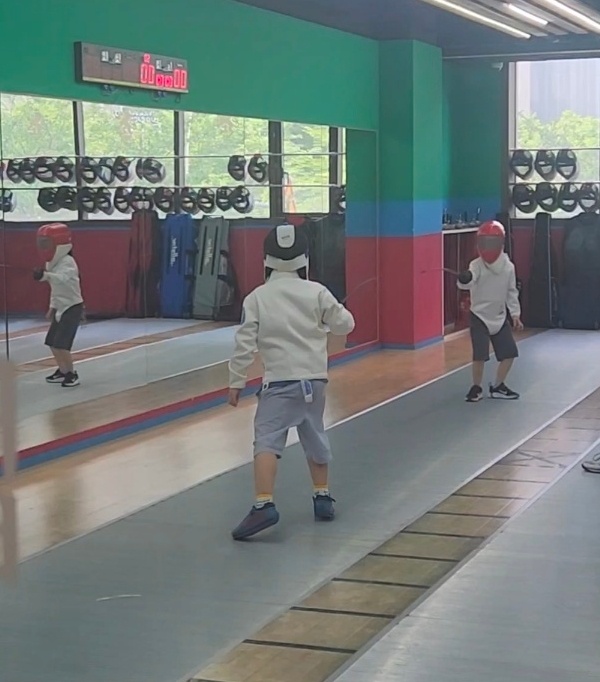 7살 딸이 펜싱장에서 마스크를 쓰고 상대와 마주 선 채로 앙가르드 자세를 잡고있다