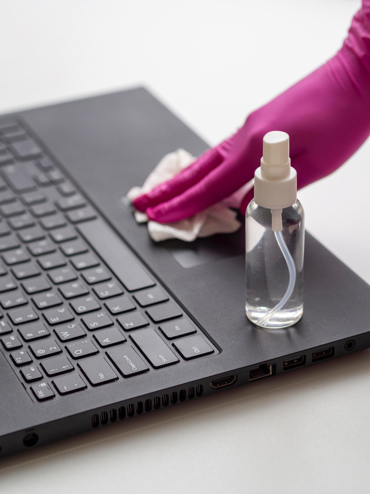 분홍색 고무장갑을 낀 손이 노트북 키보드를 극세사 천으로 닦는 모습. 옆에 투명한 스프레이 병이 놓여있음.