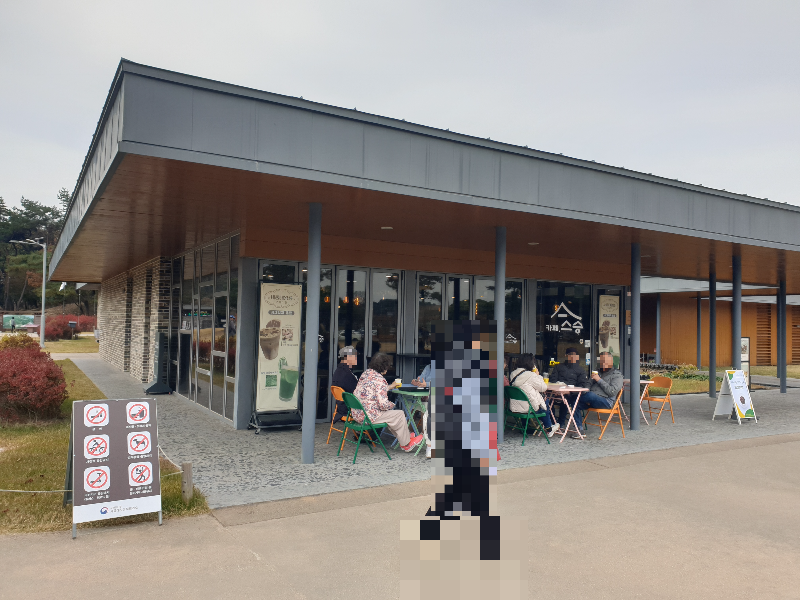 회색과 우드 그리고 유리로 구성된 카페. 야외 테이블에 많은 손님들이 앉아 있다.