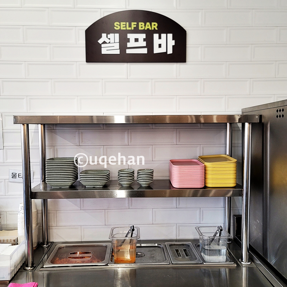 양주 칼국수 밀곳에 셀프바 사진이다. 김치와 단무지가 있고, 옆에 집게 통이 있다. 윗칸에는 앞접시들이 올려져 있다.
