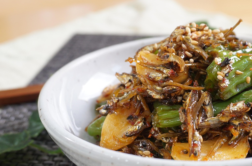 하얀 접시에 담긴 고소하고 짭조름한 멸치볶음. 마늘 슬라이스와 통깨가 뿌려져 있으며 식탁 위에 젓가락과 함께 놓인 모습