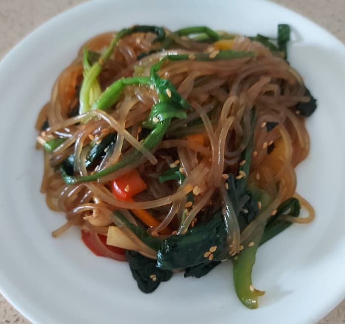 흰 접시에 담긴 잡채. 시금치, 당근, 피망, 양파 등 다양한 채소가 어우러져 있고, 참깨가 고명으로 뿌려져 있다