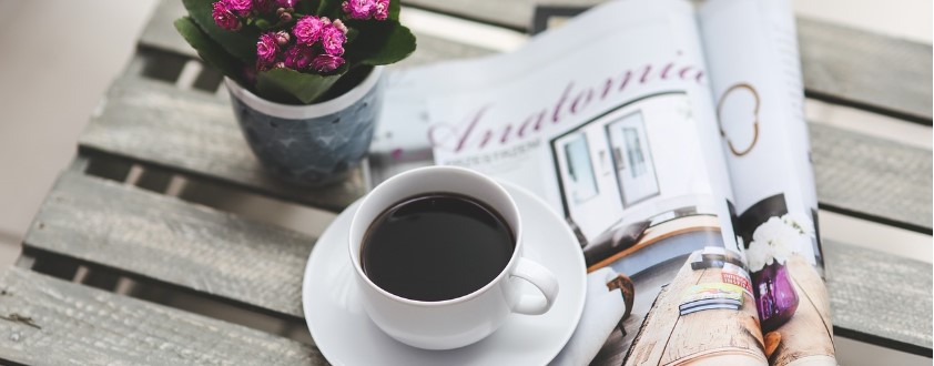 나무 테이블 위에 커피, 작은 꽃 화분, 펼쳐진 잡지책이 놓여져 있는 사진