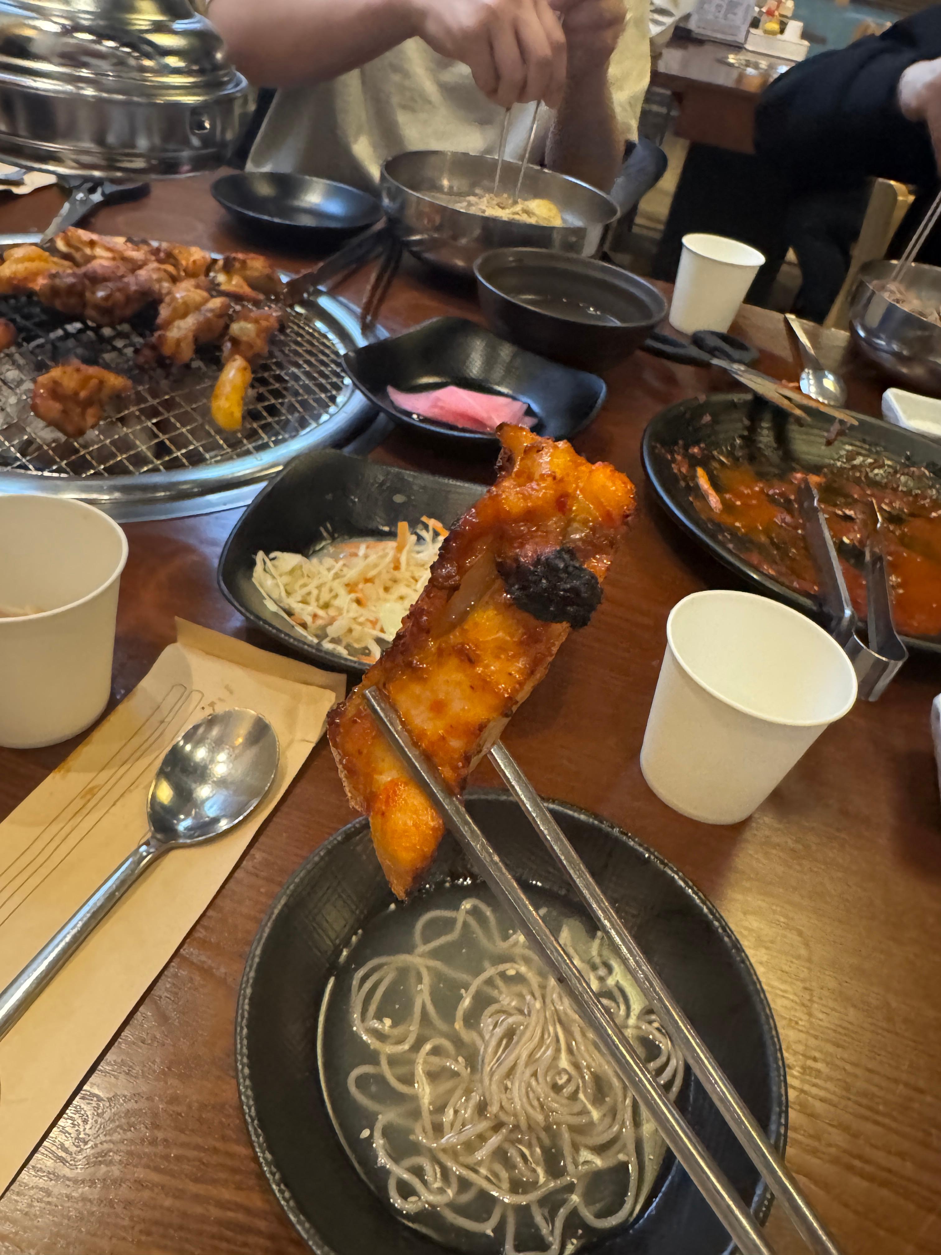 숯불 위에서 노릇하게 익은 양념닭갈비를 젓가락으로 든 클로즈업. 뒤에는 화로에 익어가는 닭갈비와 막국수 그릇들이 보인다