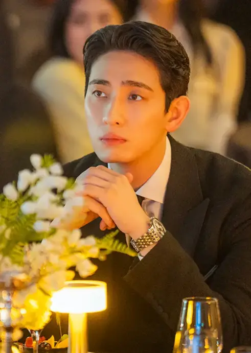 닥터슬럼프- 흰색꽃과 조명등 물잔이 놓여 테이블에 검은 쟈켓 흰색 셔츠 검은넥타이를 메고 두손을 턱에 괴고 무표정하게 있는 윤박